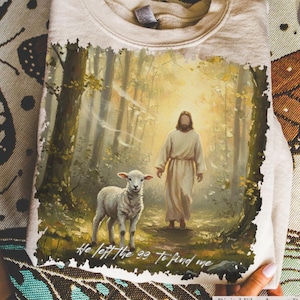 May include: Beige sweatshirt featuring a religious illustration of Jesus with a lamb in a forest setting. The artwork includes the text "He left the 99 to find me" at the bottom. The sweatshirt is held up by hands.