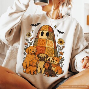 May include: Cream-colored sweatshirt featuring a colorful ghost design with a patchwork pattern, surrounded by illustrations of dogs, flowers, bats, and stars. The design has a fall or Halloween theme.