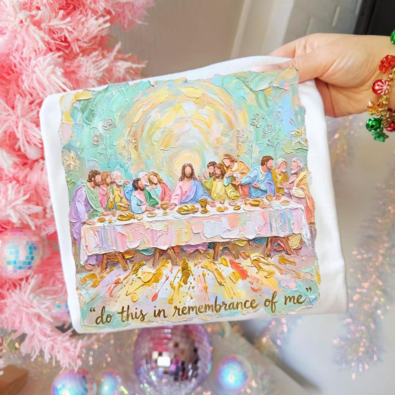 Puede incluir: Camiseta blanca con una pintura texturizada de la &Uacute;ltima Cena. La obra de arte representa a Jes&uacute;s y sus disc&iacute;pulos en una mesa, con el texto "do this in remembrance of me" debajo.