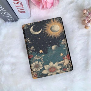 Può includere: Una custodia rettangolare con un design celeste raffigurante una luna crescente, un sole, stelle e fiori. La tavolozza dei colori comprende blu navy, oro e crema. La custodia è probabilmente per un tablet o un e-reader.