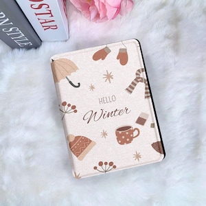 Puede incluir: Una funda de tableta beige con ilustraciones de temática invernal. El diseño incluye un paraguas marrón, mitones, una bufanda, un gorro, una taza y copos de nieve. Las palabras "HELLO Winter" están escritas en marrón.
