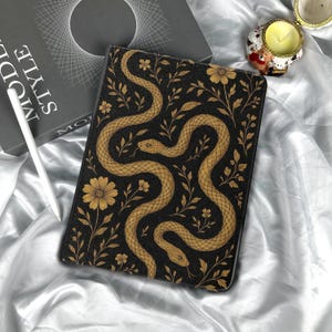 Può includere: Custodia per tablet nera con un design a serpente e fiori dorati. La custodia presenta due serpenti che si snodano tra fiori e foglie dorate dorate su sfondo nero. Sono visibili anche uno stilo bianco e un libro con la parola "STYLE".