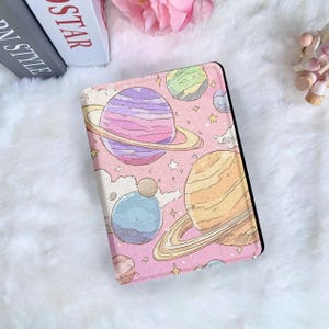 以下が含まれることがあります： ピンク色のタブレットケース。宇宙をテーマにしたデザインで、カラフルな惑星、星、雲が描かれています。惑星にはリングがあり、紫、黄色、緑、青の色合いです。ケースには黒い縁取りがあります。