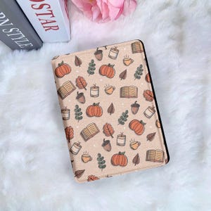 May include: A beige e-reader cover with a repeating pattern of orange pumpkins, acorns, leaves, books, candles, and coffee cups. The cover is on a white fluffy surface.