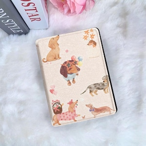 Könnte beinhalten: Eine beige Tablet-Hülle mit Aquarellillustrationen von Dackel-Hunden in verschiedenen Posen. Einige Hunde tragen Accessoires wie Sonnenbrillen, ein gestreiftes Hemd und ein Blumenkleid. Die Hülle hat einen schwarzen Rand.