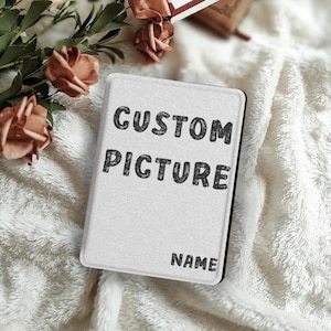 Puede incluir: Una funda rectangular blanca con las palabras "CUSTOM PICTURE" y "NAME" impresas en negro. La funda tiene un borde negro y está rodeada de una tela suave y blanca con textura y flores.