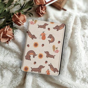 Pode incluir: Uma capa de tablet bege com um padrão repetido de dachshunds castanhos, abóboras, girassóis, bolotas e folhas de outono. A capa tem uma borda preta e está sobre um fundo macio, branco e texturizado.