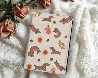 Peut inclure: Une housse de tablette beige avec un motif répété de teckels bruns, de citrouilles, de tournesols, de glands et de feuilles d'automne. L'étui a une bordure noire et est posé sur un fond doux et blanc texturé.
