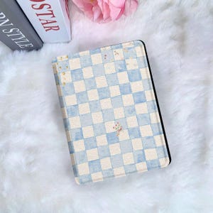 Puede incluir: Funda para tableta con un patrón de cuadros azules y blancos con un motivo floral. La funda tiene un borde negro y está sobre un fondo blanco y esponjoso. El diseño recuerda a una pintura de acuarela.
