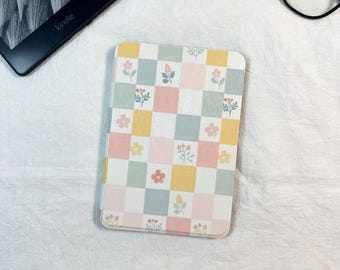 Puede incluir: Una funda rectangular para lector electrónico con un patrón de cuadros en tonos pastel suaves de rosa, amarillo y verde salvia. Cada cuadrado contiene una pequeña y delicada ilustración floral en tonos apagados, creando un motivo botánico.