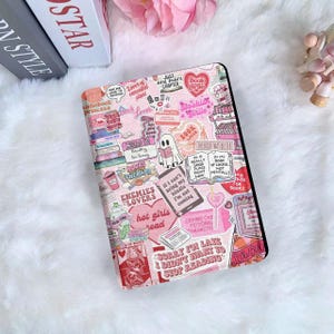 Puede incluir: Funda rosa y negra para lector de libros electrónicos con un collage de pegatinas con temática de libros. Las pegatinas incluyen frases como "Book Girlie" y "Enemies to Lovers". La funda también presenta un fantasma leyendo un libro.