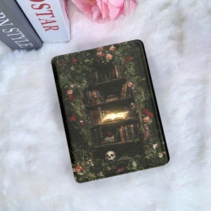 Puede incluir: Una funda rectangular para lector electrónico con un estampado oscuro y detallado de una estantería de madera cubierta de enredaderas verdes y rosas rojas. Los estantes contienen varios libros, uno de ellos brilla intensamente, y un cráneo humano descansa en la base.
