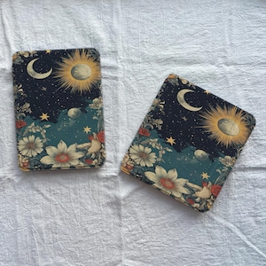 May include: Two rectangular decorative items with a celestial design. The design features a crescent moon, sun, stars, flowers, and birds against a dark blue background. The items have a vintage, artistic aesthetic.