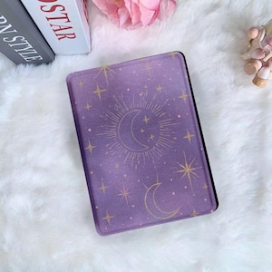 May include: A lilac e-reader cover with a celestial design featuring a crescent moon, stars, and a sunburst. The cover has a black border. The design is in gold and set against a white, fluffy background.