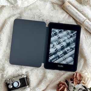 May include: A black Kindle e-reader with a gray cover, displaying a newspaper-like image. The Kindle is open, revealing the screen. A miniature camera and a rolled-up musical score are also present, along with flowers, all on a textured white surface.
