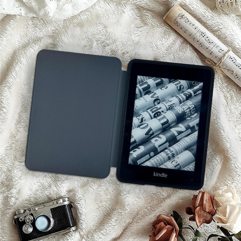 May include: A black Kindle e-reader with a gray cover, displaying a newspaper-like image. The Kindle is open, revealing the screen. A miniature camera and a rolled-up musical score are also present, along with flowers, all on a textured white surface.