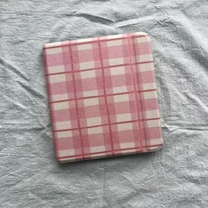 Puede incluir: Una funda para cuaderno o tableta con estampado de cuadros rosas y blancos. La funda tiene un diseño de cuadrícula con cuadrados rosas y blancos alternados. El fondo es de tela gris claro.