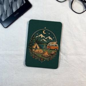 May include: A dark green e-reader case featuring a circular illustration of a camping scene. The design includes a mountain range, a crescent moon, a tent, a camper van, a campfire, and a dog resting on the ground, all framed by pine branches.