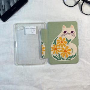 Pode incluir: Uma capa protetora para leitor eletr&oacute;nico com uma estrutura de pl&aacute;stico transparente &agrave; esquerda e uma capa verde-s&aacute;lvia &agrave; direita. A capa apresenta uma ilustra&ccedil;&atilde;o de um gato creme decorado com flores amarelas e folhas verdes.