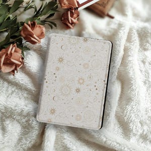 Pode incluir: Uma capa de tablet creme com um design celestial de sóis, luas, estrelas e nuvens douradas. A capa tem uma borda preta e está sobre uma superfície branca e fofa. Flores decorativas no fundo.
