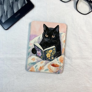 Puede incluir: Ilustración en acuarela de un gato negro leyendo un libro con diseños florales. El gato está en una cama con una manta floral y una taza de té a su lado. Un lector Kindle es visible en el fondo.