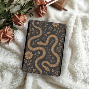 Puede incluir: Una funda negra con un diseño de serpiente y flores doradas. La funda tiene un fondo oscuro con serpientes, flores y hojas doradas. La funda rectangular es probablemente para una tableta o un lector electrónico.