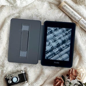 May include: An open gray Kindle e-reader with a newspaper-like display, next to a matching gray case. A vintage-style camera and a rolled-up musical score are also in the image, along with a few roses.