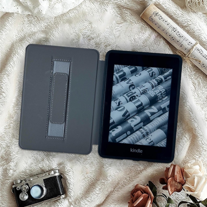 May include: An open gray Kindle e-reader with a newspaper-like display, next to a matching gray case. A vintage-style camera and a rolled-up musical score are also in the image, along with a few roses.