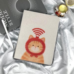 May include: A tablet case featuring a cartoon orange tabby cat wearing a red hat with a heart and a Wi-Fi symbol. The case has a textured, off-white background. A white stylus and a book with the words "MODE STYLE" are visible.