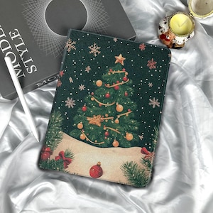 以下が含まれることがあります： 赤と金色のオーナメント、星、雪の結晶が飾られたクリスマスツリーのデザインの濃い緑色のタブレットケース。ケースには雪と赤いリボンが描かれた冬のシーンがあります。ケースは白いサテンの表面にあります。
