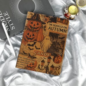 May include: A Halloween-themed journal with orange pumpkins, a ghost, and bats on a brown background. The text "IM HOOKED ON AUTUMN!" and "CANDY" are visible. The journal is on a white satin surface.