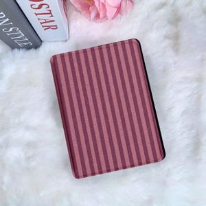 Peut inclure: Un étui rectangulaire à rayures bordeaux et rose foncé, probablement pour un lecteur électronique ou une tablette, repose sur une surface blanche et duveteuse. Le fond comprend des livres et une fleur rose.