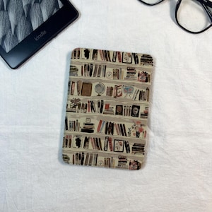 May include: A rectangular e-reader cover featuring a repeating pattern of bookshelves filled with various books, a globe, a clock, and floral arrangements in muted tones of beige, brown, black, and soft pink, with a black Kindle device visible nearby.