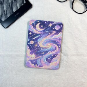May include: A rectangular e-reader skin featuring a vibrant cosmic illustration of a swirling galaxy, glowing stars, planets, and a crescent moon in shades of purple, blue, pink, and yellow against a deep space background.