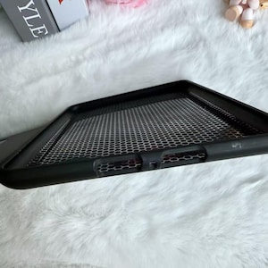 May include: Black phone case with a honeycomb pattern. The case has a raised edge and a textured surface. The background is a soft, white, fluffy material.