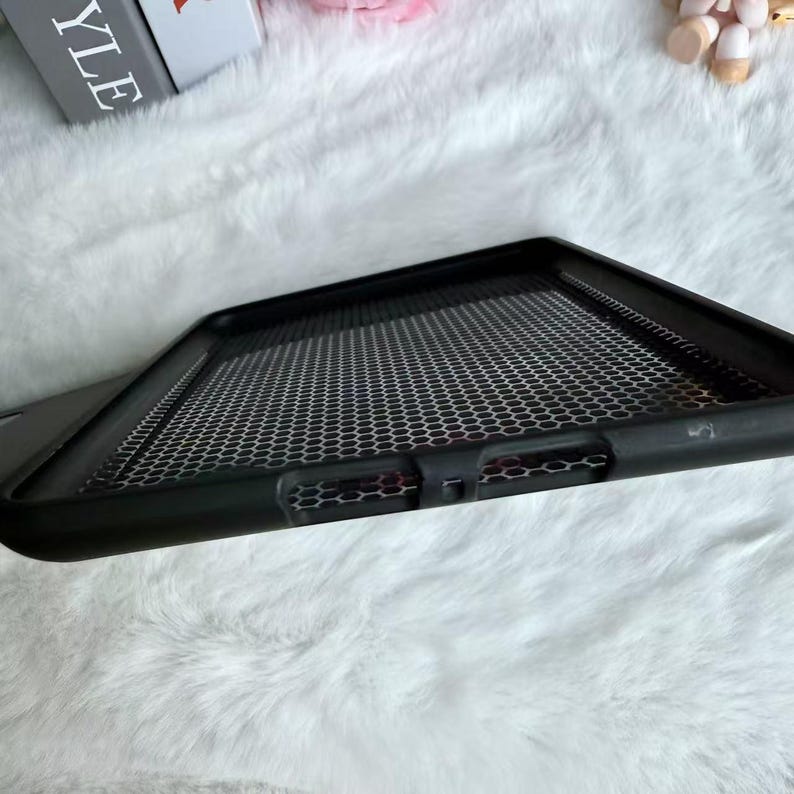 May include: Black phone case with a honeycomb pattern. The case has a raised edge and a textured surface. The background is a soft, white, fluffy material.