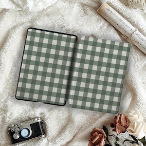 May include: An open e-reader case with a green and white gingham pattern. The case is black and sits on a white, fluffy surface. A vintage camera, sheet music, and flowers are also in the image.