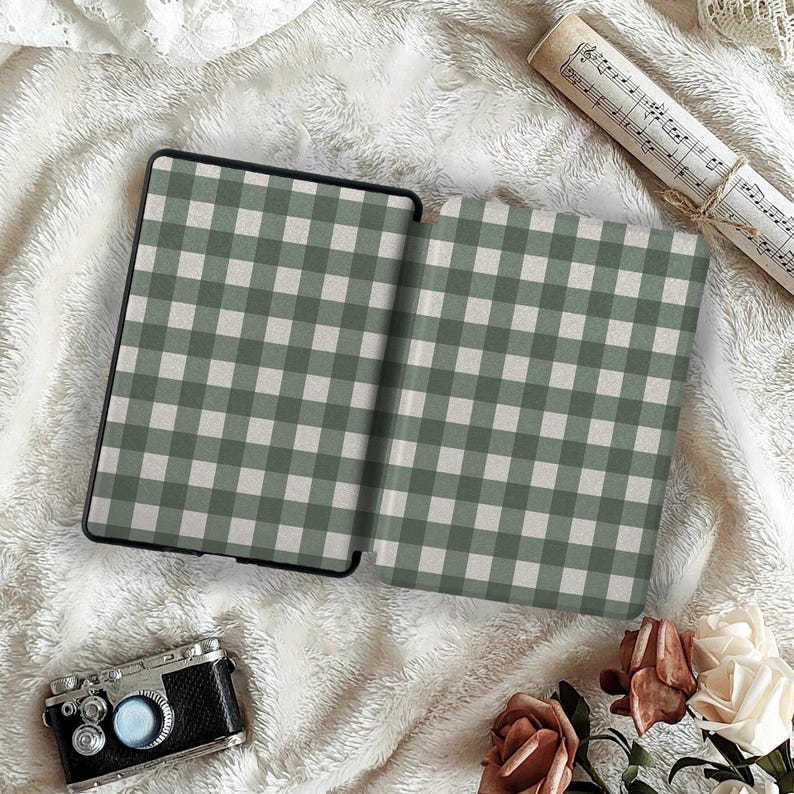 May include: An open e-reader case with a green and white gingham pattern. The case is black and sits on a white, fluffy surface. A vintage camera, sheet music, and flowers are also in the image.