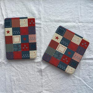 May include: Two square, padded items with a patchwork design in red, white, and blue. The design includes stars, stripes, and squares. The items are likely coasters or placemats, with a patriotic theme.