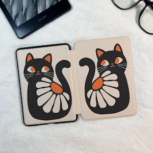 Pode incluir: Uma capa dobr&aacute;vel para leitor de e-books com fundo creme e duas ilustra&ccedil;&otilde;es de gatos pretos. Cada gato tem orelhas laranja, olhos laranja e um padr&atilde;o de flor branca semelhante a uma margarida na lateral. A capa est&aacute; aberta.