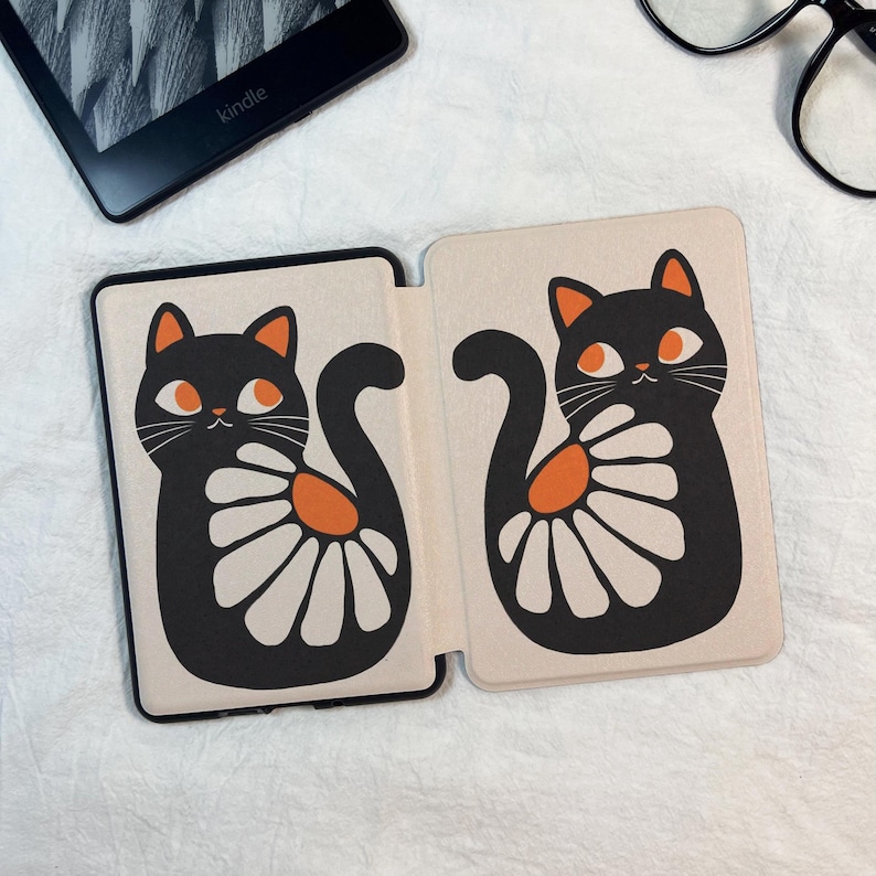 Pode incluir: Uma capa dobr&aacute;vel para leitor de e-books com fundo creme e duas ilustra&ccedil;&otilde;es de gatos pretos. Cada gato tem orelhas laranja, olhos laranja e um padr&atilde;o de flor branca semelhante a uma margarida na lateral. A capa est&aacute; aberta.