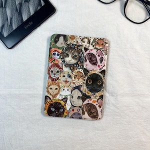 Op de afbeelding: Een rechthoekige Kindle-hoes met een patroon van kattengezichten met capuchons. De katten hebben verschillende kleuren, waaronder zwart, grijs en oranje. De hoes lijkt gemaakt te zijn van een glad materiaal.
