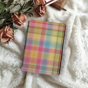 以下が含まれることがあります： 黒い縁取りのカラフルなチェック柄ノート。チェック柄はピンク、黄色、青、緑の正方形で構成されています。ノートは白くふわふわした表面に置かれ、背景には桃色のバラがいくつかあります。