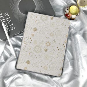 May include: A light-colored tablet case with a celestial design featuring suns, moons, stars, and constellations in gold. The case is lying on a satin surface next to a book and a white stylus.