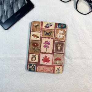 Puede incluir: Una funda Kindle con un collage de diseños de sellos postales vintage con flores, insectos y un búho. La funda es en tonos marrón, beige y rojo, con el texto "Enjoy Every Moment".