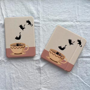 May include: Two beige tablet cases with a pink bottom section. Each case features black cat silhouettes seemingly playing around a coffee cup illustration. The cases are on a white, textured surface.