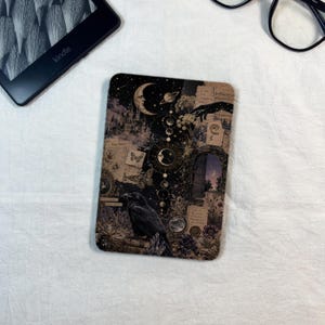 May include: A rectangular e-reader skin featuring a dark collage design with a black raven, a crescent moon, celestial phases, a castle silhouette, vintage paper scraps, and floral elements in shades of black, cream, and muted purple tones.