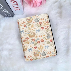 May include: A cream-colored tablet case featuring a whimsical pattern of geese with blue bows, baskets of flowers, mushrooms, hearts, and a teapot. The case has a black border and is set against a fluffy white background.