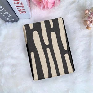 Puede incluir: Una funda rectangular con un patrón abstracto en negro y beige. El diseño presenta formas beige verticales sobre un fondo negro. La funda está sobre una superficie blanca y esponjosa.