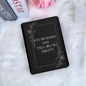 Könnte beinhalten: Schwarzer rechteckiger Buchumschlag mit dem weißen Text "BUY ME BOOKS AND TELL ME I'M PRETTY". Florale Linienkunst akzentuiert den Text. Der Umschlag liegt auf einer weißen, flauschigen Oberfläche.